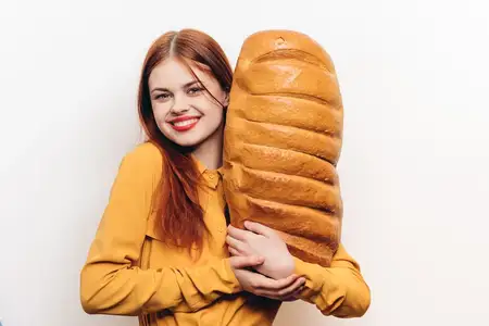 Счастливая женщина держит в руке большую буханку хлеба в вертикальном положении, улыбается, позирует как модель
