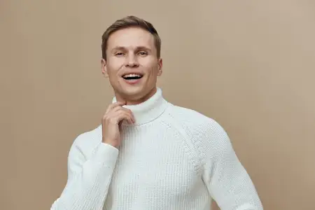 Веселый мужчина с дружелюбной улыбкой, одетый в уютный белый свитер, позирует на фоне однотонного бежевого фона