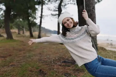 Улыбающаяся женщина на качелях в зимнем пейзаже у озера, в уютном свитере и шапке-бини, наслаждающаяся свежим воздухом и активным отдыхом среди деревьев и открытого пространства