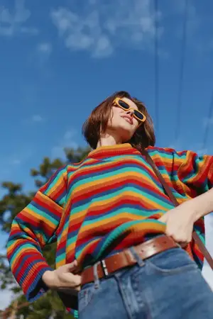 Молодая взрослая женщина в радужном полосатом свитере и ретро-солнцезащитных очках на фоне голубого неба, повседневный наряд, руки на бедрах, уверенно улыбающаяся в солнечном городском образе жизни…