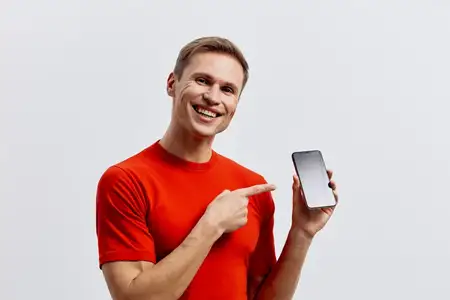Улыбающийся мужчина в оранжевой футболке держит смартфон и указывает пальцем на экран на белом фоне