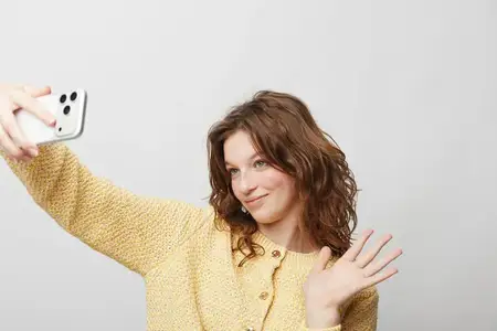 Улыбающаяся кудрявая женщина в желтом свитере держит смартфон для селфи и машет рукой