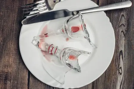 Разбитый бокал для вина, нож и вилка, осколки на деревянном столе