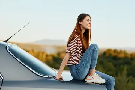 Женщина-водитель сидит на багажнике автомобиля и улыбается, любуясь красивым видом осенней природы и гор