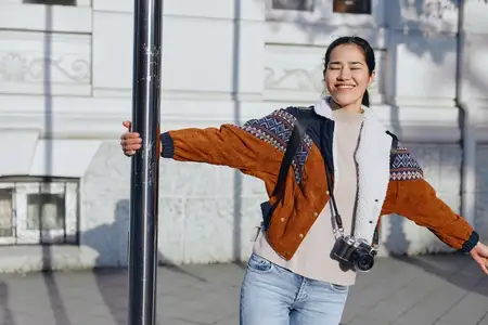 Городская уличная сцена: улыбающаяся женщина с фотоаппаратом на шее, в джинсах и объемном кардигане, позирует у фонарного столба
