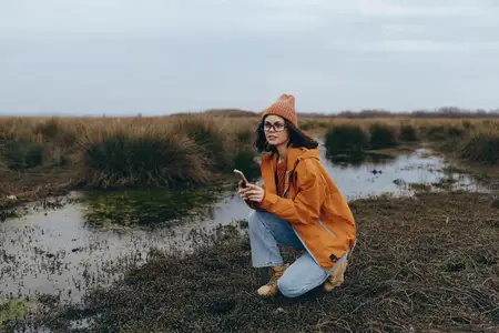Фотография женщины на отдыхе на природе: она стоит на коленях у реки осенью, в оранжевом пальто и вязаной шапке, очках