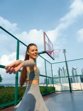 Счастливая молодая женщина в спортивной одежде играет на открытой баскетбольной площадке на фоне яркого неба