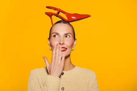 Улыбающаяся женщина с красными туфлями на высоком каблуке, балансирующими на голове, в серьгах и бежевом свитере на жёлтом фоне