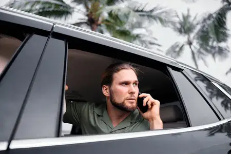 Крупный план мужчины, который смотрит из окна автомобиля и разговаривает по телефону на фоне тропической природы