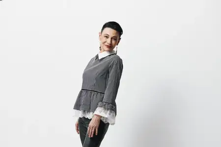 Женщина в серой рубашке и черных джинсах стоит, положив руки на пояс, на фоне белого фона
