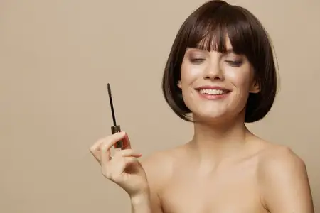 Молодая женщина красит ресницы, обнажённые плечи, изолированный фон