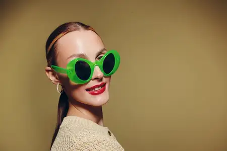 Уверенная молодая женщина в зеленых круглых солнцезащитных очках и бежевом свитере позирует на бежевом фоне