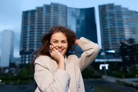 Счастливая молодая женщина в бежевом пальто уверенно позирует на улице на фоне современных городских зданий и облачного неба, излучая позитивные эмоции и чувство силы
