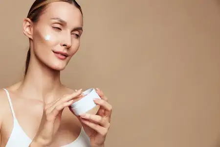 Улыбающаяся женщина с чистой кожей использует крем для лица на щеке, держа косметическую банку