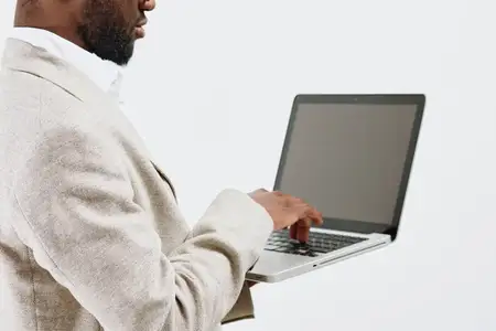 Профессиональный мужчина в бежевом свитере работает с открытым ноутбуком, стоит в помещении на однотонном фоне, сосредоточен на цифровом устройстве