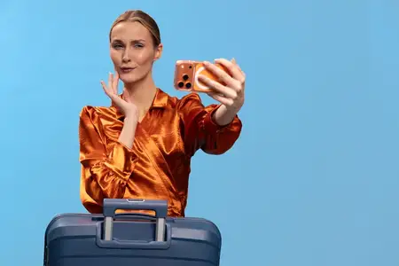 Модная женщина делает селфи на оранжевый смартфон, держа синий чемодан в студии с голубым фоном