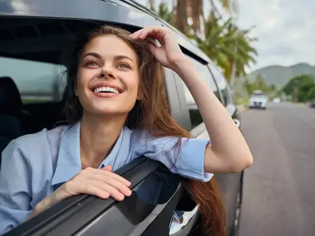 Улыбающаяся женщина выглядывает из окна автомобиля на фоне пальмовых деревьев, передавая беззаботную и тропическую атмосферу