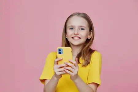 Молодая девушка в ярко-желтой одежде улыбается и держит смартфон на фоне жизнерадостного розового фона, излучая позитив и юношескую энергию