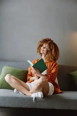 Молодая женщина с вьющимися волосами с удовольствием читает книгу на зелёном диване, одетая в повседневную одежду и выражающая радость
