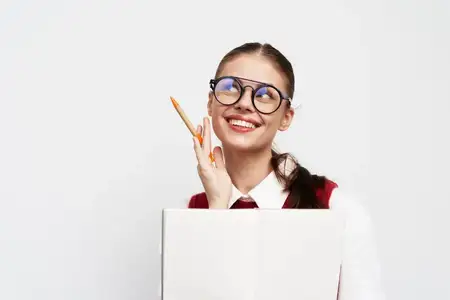 Весёлая молодая женщина в очках, одетая в белую рубашку и красный жилет, держит большую тетрадь и карандаш