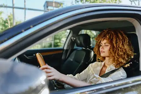 Молодая женщина с вьющимися волосами ведет черный автомобиль, пристегнувшись ремнем безопасности и надев легкую блузу