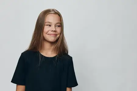Улыбающаяся юная девушка с прямыми волосами, одетая в повседневную чёрную футболку, позирует на светлом однотонном фоне, глядя в сторону с радостным лицом и расслабленной позой