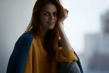 Миловидная женщина в желтом свитере прячется за одеялом у окна в уютном домашнем интерьере