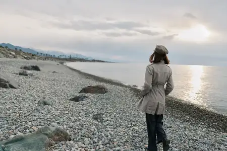 Одинокая женщина в осенней одежде идет по галечному пляжу у спокойной воды на закате