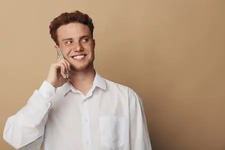 Счастливый рыжеволосый мужчина в белой рубашке прижимает смартфон к уху и с улыбкой смотрит в сторону на бежевом фоне