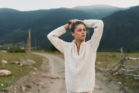 На пыльной проселочной дороге на фоне гор женщина в белом вязаном свитере отводит взгляд в сторону, пребывая в спокойном, задумчивом настроении, а естественный свет подчеркивает безмятежный сельский…