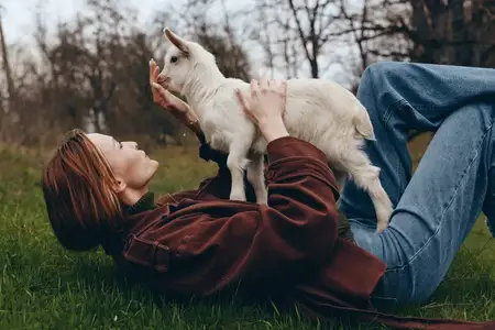 Молодая женщина в коричневой куртке и джинсах лежит на траве, держа и взаимодействуя с белым козлёнком в мирной обстановке на открытом воздухе