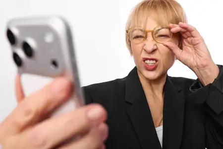Пожилая женщина в очках со смартфоном делает селфи, но выражает стресс и напряжение глаз, прикасаясь рукой к глазу во время просмотра экрана