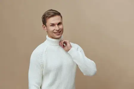 Улыбающийся мужчина в белом вязаном свитере смотрит в камеру с дружелюбным выражением лица, позируя на фоне однотонного бежевого фона