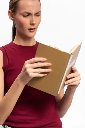 Растерянная молодая женщина в красном топе без рукавов читает открытую книгу с сосредоточенным выражением лица