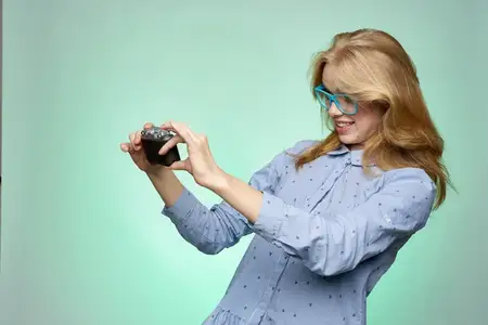 Женщина-блондинка, фотограф в очках, с фотоаппаратом в руках на зелёном фоне, в синей рубашке