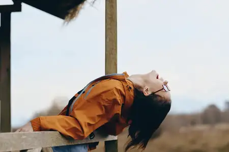 Женщина на отдыхе на природе: опирается на деревянный забор, оранжевая куртка, длинные волосы развеваются на ветру, расслабленный взгляд вверх, спокойная улыбка, дневной портрет, фон неба со…