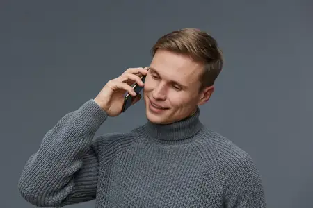 Улыбающийся молодой светлокожий мужчина в сером вязаном свитере держит мобильный телефон возле уха, глядя вниз, изолирован на темно-сером фоне