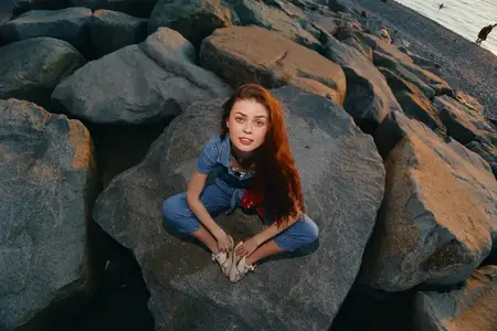 Молодая женщина в стиле лайфстайл, одетая в современный бохо-вестерн деним стритстайл, непринуждённо сидит на больших камнях у пляжа во время заката, снятая в тёплых плёночных цветовых тонах