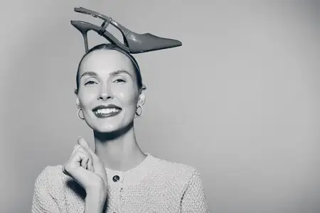 Счастливая молодая светлокожая женщина европеоидной расы балансирует туфлёй на высоком каблуке на голове, одетая в уютный свитер