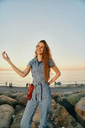 Беззаботная женщина в стиле жизни, одетая в современный бохо-вестерн джинсовый стритстайл, стоит на камнях у океана на закате, улыбаясь с естественной радостью, с эффектом винтажной плёнки