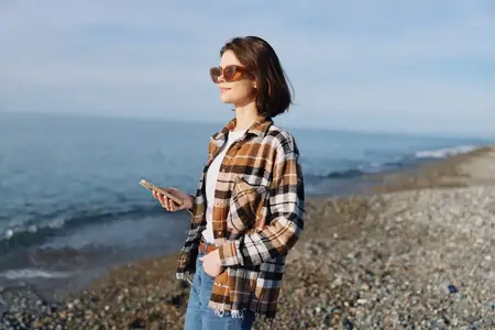 Повседневная женщина у моря со смартфоном, ветер в волосах и солнцезащитные очки, джинсовая куртка, расслабленная поза на берегу для образа жизни, путешествий, хорошего самочувствия и исследования…