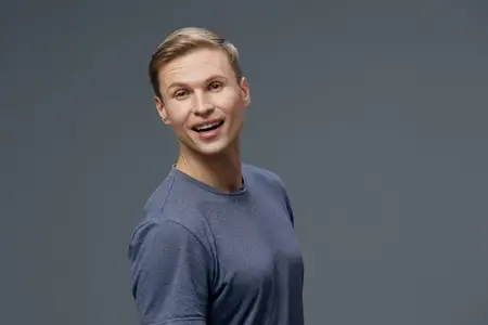 Счастливый молодой человек с короткими светлыми волосами, одетый в удобный синий свитер, широко улыбается с радостным выражением лица, стоя на фоне однотонного серого фона