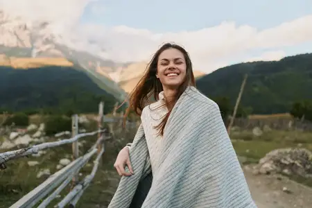 Жизнерадостная женщина, укутанная в уютное одеяло, стоит на улице на фоне гор; расслабленная поза, улыбающееся лицо, естественная обстановка, повседневный образ жизни, свежий воздух