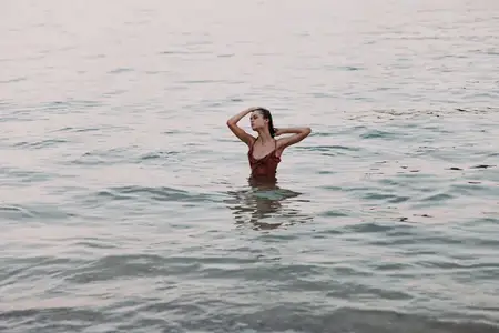 Молодая женщина в красном купальнике стоит в спокойной морской воде во время заката, наслаждаясь безмятежной атмосферой и мягким теплым светом вечера