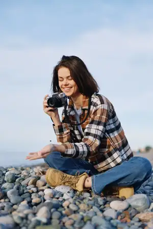Повседневный фотограф наслаждается природой, фотоаппарат наготове на галечном пляже, женщина в ботинках и клетчатой рубашке позирует с радостным выражением лица, документируя детали и текстуры…