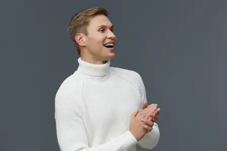 Счастливая женщина с короткими светлыми волосами, одетая в уютный белый вязаный свитер с высоким воротником, ярко улыбается и смотрит в сторону, празднуя или выражая радость на фоне однотонного…