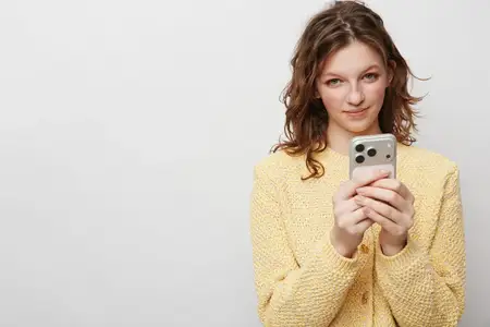 Улыбающаяся женщина в желтом вязаном свитере пользуется смартфоном, глядя прямо в камеру
