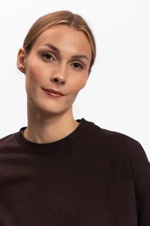 Молодая женщина с blond-волосами в темно-коричневом топе позирует с безмятежным выражением лица на белом фоне