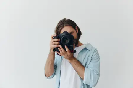 Мужчина-хипстер, фотограф, в студии на фоне белого фона смотрит в видоискатель камеры и делает снимки при естественном свете из окна
