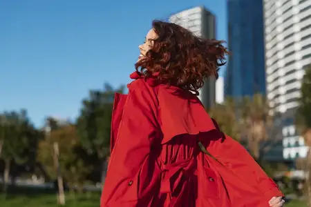 Женщина в красном пальто идет по городскому пейзажу, ветер развевает ее волосы и пальто, на заднем плане небоскребы, запечатлен непринужденный момент современной моды и свободы в городе